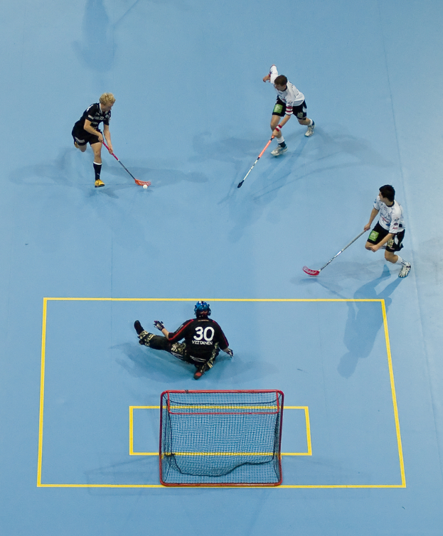 Drei Personen in Sportkleidung spielen Hockey, wobei eine Person rechts mit einem Hockeystock den Ball schlägt und eine Person hinter einem Netz auf dem Boden sitzt.