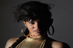 Eine Frau in einem goldenen Kleid und großen Creolen steht selbstbewusst vor einer Wand, ihre Haare sind zu einem sleeken Bob gestylt und sie schaut direkt in die Kamera mit einem entschlossenen Ausdruck.