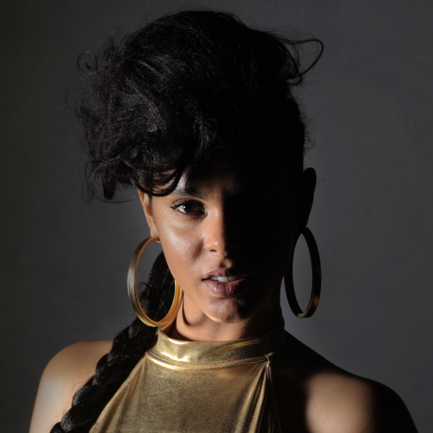 Eine Frau in einem goldenen Kleid und großen Creolen steht selbstbewusst vor einer Wand, ihre Haare sind zu einem sleeken Bob gestylt und sie schaut direkt in die Kamera mit einem entschlossenen Ausdruck.