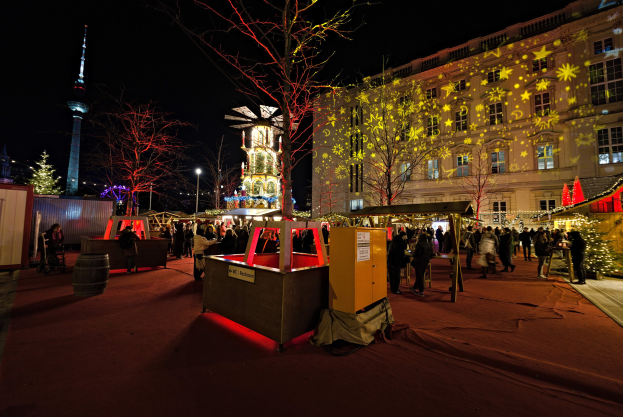Ein lebendiger Weihnachtsmarkt in Berlin, Deutschland, mit Menschen um beleuchtete Stände, Bäume, Gebäude, Laternenmasten und einen Turm unter einem dunklen Himmel.