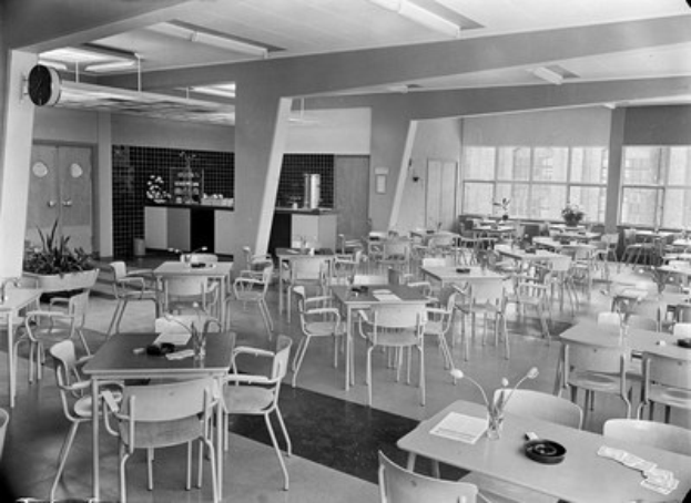 Schwarz-weiß-Foto einer Cafeteria mit Tischen und Stühlen, Tellern, Besteck, Gläsern und Vasen auf den Tischen und Türen und Fenstern im Hintergrund unter Deckenlampen.