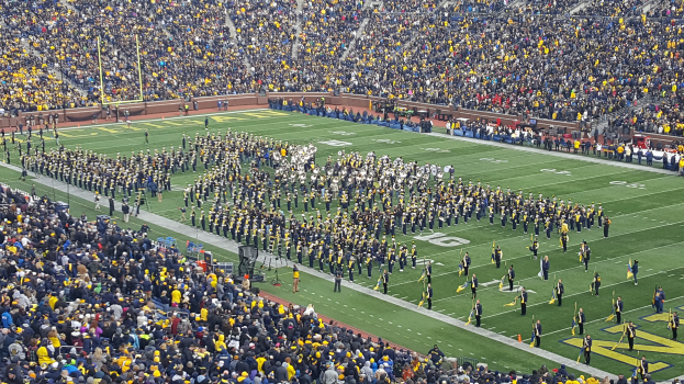 Eine große Menschenmenge in einem Stadion bei einem Fußballspiel, wobei einige stehen und Fahnen halten, andere sitzen und Pfosten und Geländer im Hintergrund zu sehen sind.