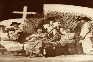 Schwarzes und weißes Foto einer Mariachi-Band, die vor einer Kirche mit einem Kreuz an der Wand dahinter sitzt.