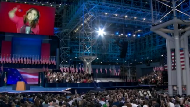 Eine große Menschenmenge sitzt vor einer Bühne mit einem Rednerpult, Mikrofon, Flaggen, einem Bildschirm mit dem Bild einer Frau und Beleuchtungsausrüstung und wartet auf eine Präsidentschaftsdiskussion.