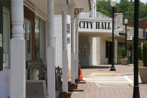 Ein Rathaus umgeben von Gebäuden, Straßenmästen, Straßenlaternen, Namensschildern, Kraftfahrzeugen, Absperrgittern, Stühlen, Topfpflanzen, Bäumen und einem bewölkten Himmel.