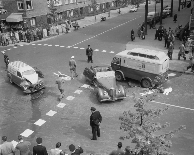 Schwarz-weiß-Szene eines Autounfalls an einer Straßenkreuzung mit mehreren Fahrzeugen, darunter ein Lieferwagen, und Passanten; Gebäude, Bäume, Pfosten und Texttafeln im Hintergrund.