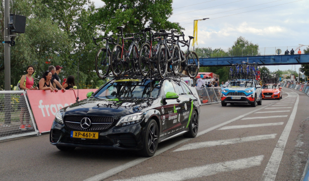 Ein Mercedes-Benz C63 AMG 4MATIC+ mit Fahrrädern auf dem Dach fährt eine Straße entlang, umgeben von Menschen, einem Zaun, einer Fahne, einem Straßenpfahl, einem Schild, einer Brücke, Bäumen, Drähten und einem bewölkten Himmel.