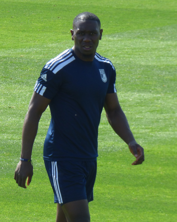 Ein Mann in einem blauen T-Shirt und Shorts geht selbstbewusst über ein grünes Feld und sieht entschlossen aus.