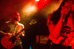 Eine Frau singt in ein Mikrofon auf der Bühne, während ein Mann Gitarre spielt.