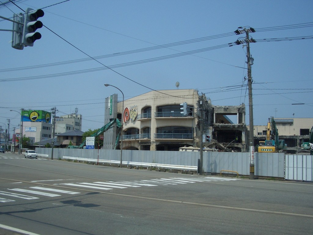 Stadtstraße mit Fahrzeugen, Strommasten, Laternen, einer Ampel, Gebäuden, Bäumen und einem teilweise abgerissenen Gebäude im Vordergrund.
