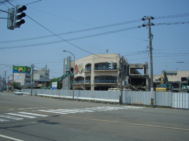 Stadtstraße mit Fahrzeugen, Strommasten, Laternen, einer Ampel, Gebäuden, Bäumen und einem teilweise abgerissenen Gebäude im Vordergrund.