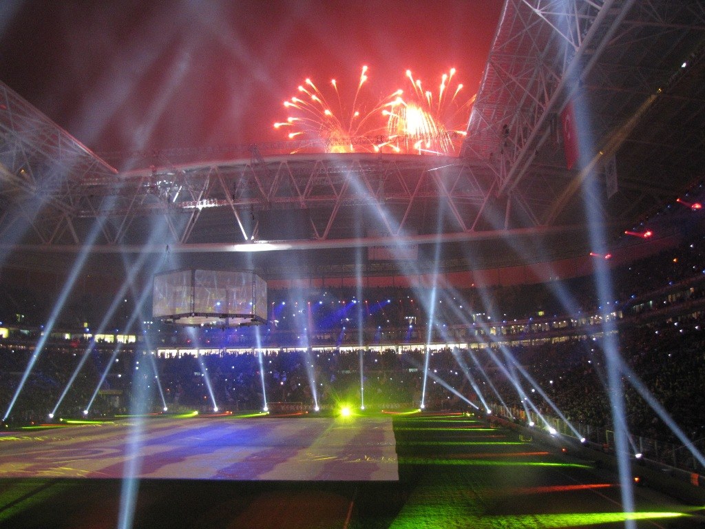 Das Olympische Stadion in London, England, bei Nacht mit hellen Lichtern und Feuerwerken erleuchtet, gefüllt mit Menschen und einem großen Bildschirm im Vordergrund.