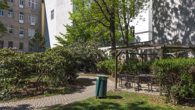 Ein Park mit einem grünen Müllcontainer in der Mitte, umgeben von Gras, Pflanzen, Bäumen und Gebäuden mit Fenstern, mit verstreuten Stühlen und Tischen und einer Hütte im Hintergrund.