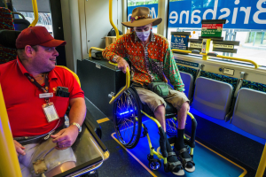 Zwei Männer im Rollstuhl im Bus, einer trägt ein rotes T-Shirt und eine rote Mütze, der andere ein orangefarbenes Hemd und einen braunen Hut, beide tragen Masken und Ausweise, mit gelben Stangen und Glasfenstern, die Bäume, Gebäude und den Himmel zeigen.