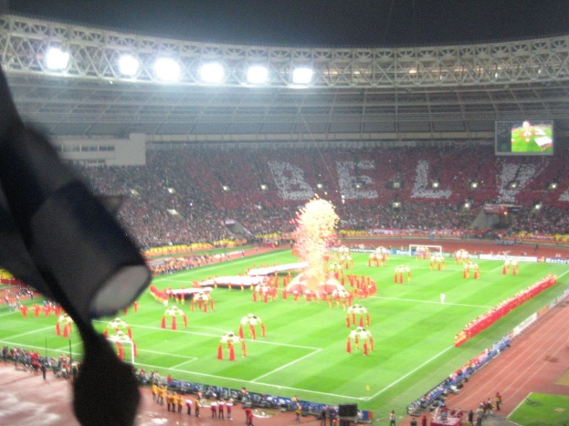 Ein Stadion mit Flutlicht, Eisengittern, Zuschauern, stehenden Individuen, Werbetafeln, Anzeigetafeln, Überwachungskameras, Fahnen und Dekorationen.