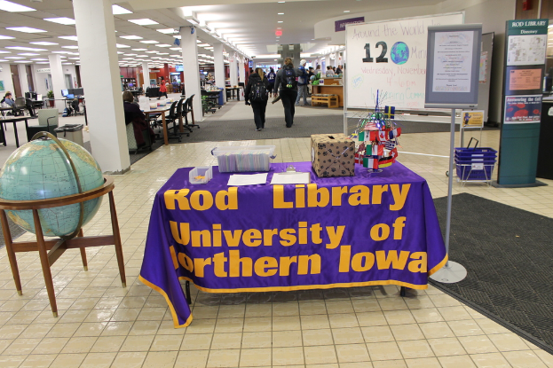 Eine Messestellung mit einem Tisch, auf dem verschiedene Landesflaggen, eine Kiste, Papiere und ein Banner sowie links daneben eine Weltkugel und rechts ein Anschlagbrett mit einigen Menschen in der N√§he und mehreren Tischen und Stühlen zu sehen sind.