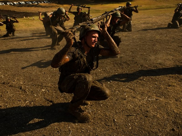 Eine Gruppe von Männern in Militäruniformen, Helmen und mit Gewehren in der Hand läuft über ein Feld mit Bäumen und Gras im Hintergrund und Text am unteren Rand des Bildes.