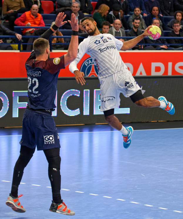 Zwei Männer spielen Handball auf einem Platz, einer springt und hält den Ball, mit Zuschauern und einer Anzeigetafel im Hintergrund.