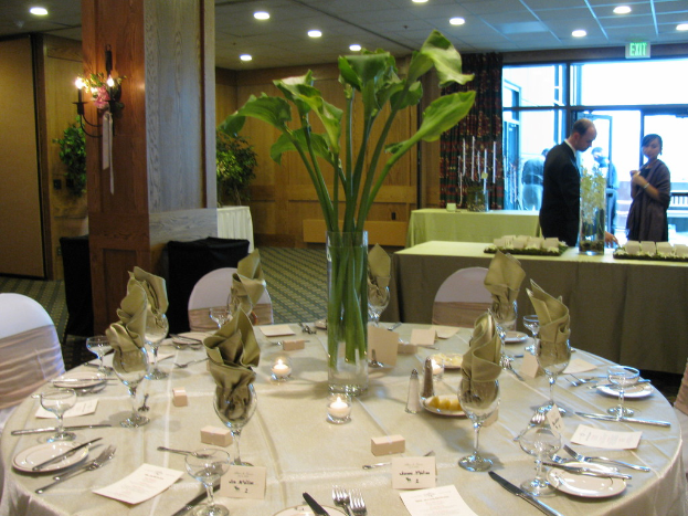 Ein Tisch mit einer weißen Decke, Tellern, Besteck, einem Glas und Baumzweigen, umgeben von Stühlen, mit einem grünen Tisch dahinter, einem Mann in einem schwarzen Blazer, einem stehenden Mädchen, einer Tür mit der Aufschrift 'Ausgang' und zwei Männern dahinter.