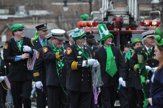 Eine Gruppe von Männern in Uniform marschiert in einem St. Patrick's Day Umzug, hält Perlen und hat ein Feuerwehrauto und Gebäude im Hintergrund.