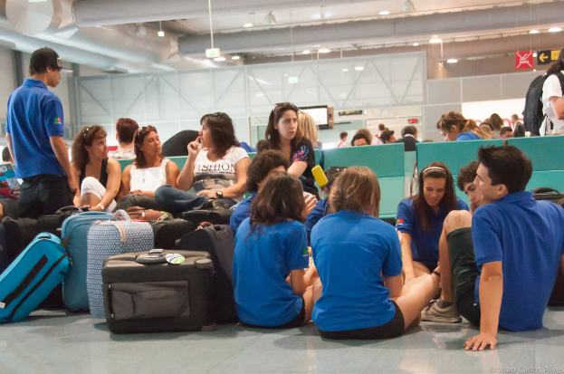 Ein belebter Flughafen mit vielen Menschen, die auf ihre Flüge warten, Gepäck zu links und gläserne Türen mit Richtungsschildern im Hintergrund.