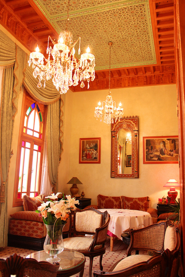 Ein Wohnzimmer mit einem Tisch und Blumentopf im Vordergrund, umgeben von Stühlen, Sofas und einem Tisch mit einer weißen Decke im Hintergrund, gerahmte Bilder an der Wand, von der Decke hängende Lampen und Vorhänge mit einem Fenster links.