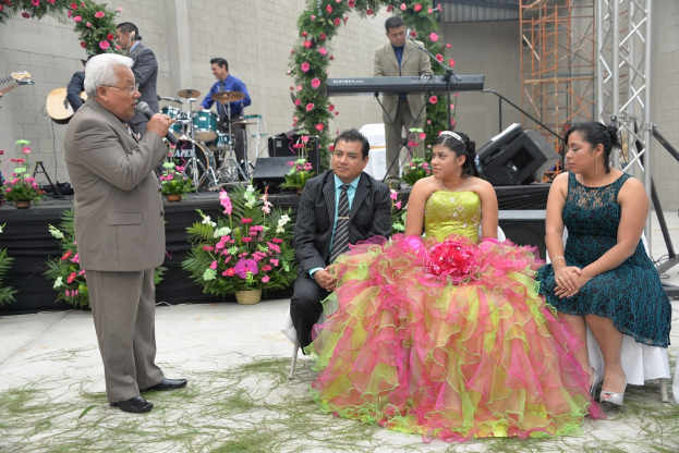 Ein Mann steht und spricht in ein Mikrofon, während andere sitzen und ein Orchester im Hintergrund spielt.