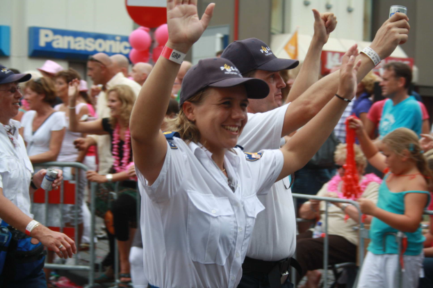 Eine Gruppe von Menschen, einige mit Mützen und Dosen haltend, steht und sitzt vor einer Menge, mit Geländern, Ballons und einem Gebäude mit Namensschildern im Hintergrund.