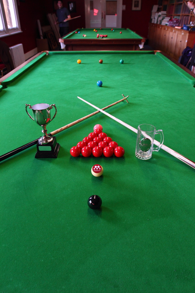 Eine Trophäe, ein Glasbecher und mehrere Kugeln sind auf einem Billardtisch angeordnet.