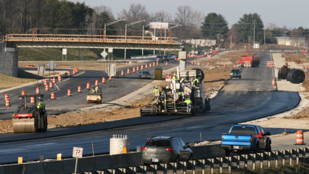 Baustelle mit Fahrzeugen auf der Straße, Verkehrskegel, Schilder, Pfosten, eine Brücke, Bäume und einen klaren blauen Himmel.