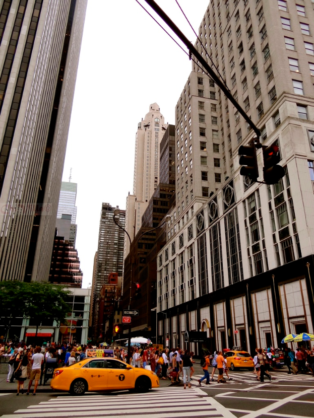 Eine Stadtstraße mit Gebäuden, Ampeln, Fenstern, wenigen Passanten, Autos und Himmel.