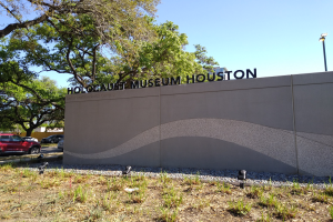 Außenansicht des Holocaust-Museums Houston mit einer Gedenkwand, Steinen, Grünfläche, Bäumen, Straßenverkehr, einem Straßenschild.