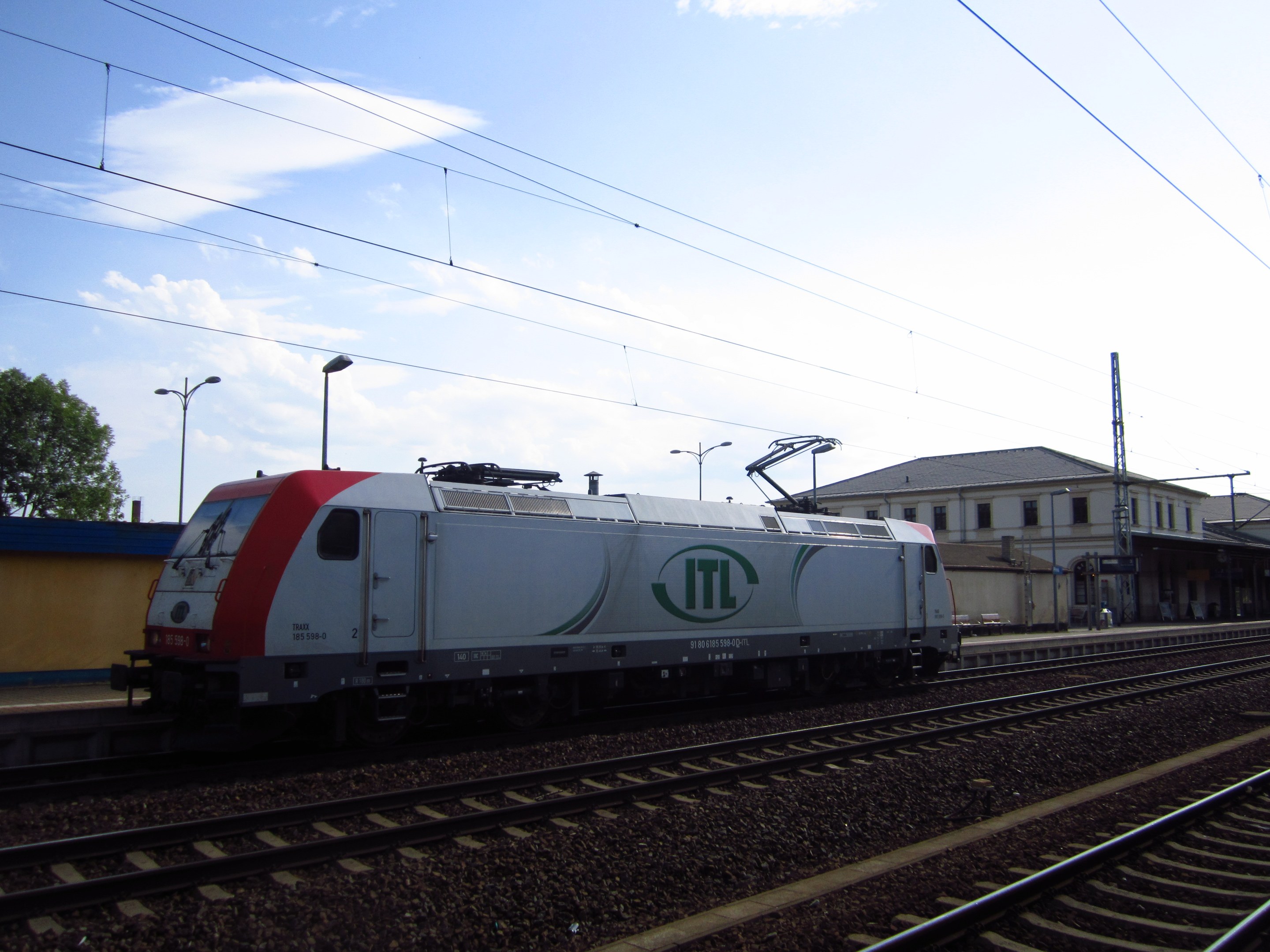 Ein diesel-elektrischer Zug fährt auf Schienen neben einem Gebäude, mit elektrischer Infrastruktur, Straßenmöblierung, Bäumen und einem bewölkten Himmel im Hintergrund.