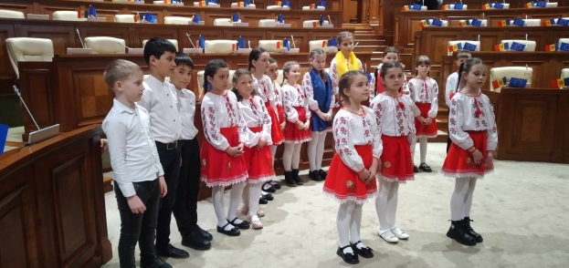 Eine Gruppe von Kindern in weißen und roten Kleidern steht zusammen in einem Raum, mit einem Podium und Mikrofon links und Stühlen und Tischen mit verschiedenen Gegenständen im Hintergrund, tragend Fahnen, die die Teilnahme an einer Nationalversammlung anzeigen.
