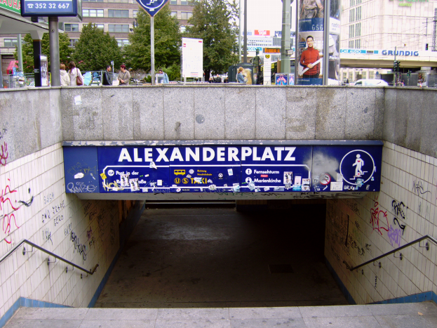 Eine U-Bahn-Station mit Graffiti an den Wänden und Treppen, Geländern auf beiden Seiten und einer Tafel mit Text und Bildern, mit Gebäuden, Bäumen, Pfählen, Menschen, Fahrzeugen und anderen Objekten im Hintergrund unter einem klaren blauen Himmel.
