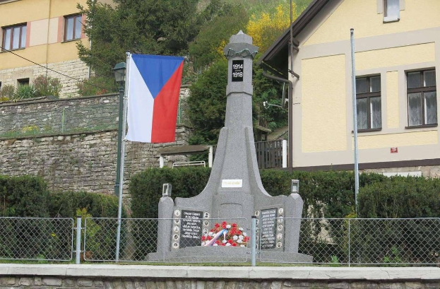 Ein Denkmal mit einer Flagge darauf steht vor einem Gebäude, umgeben von einem Zaun, Pflanzen, Bäumen und einer Wand mit eingravierten Text.