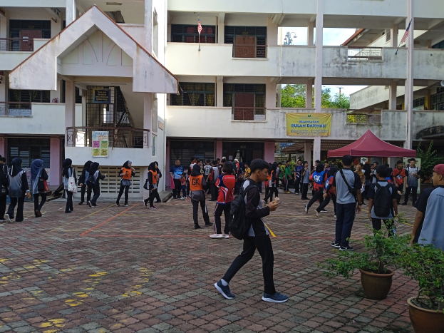 Eine Gruppe von Menschen, einige mit Taschen, geht vor einem Gebäude mit Fenstern, Geländern, Säulen, Bannern, Fahnen, einem Zelt, Topfpflanzen und Bäumen unter einem bewölkten Himmel.