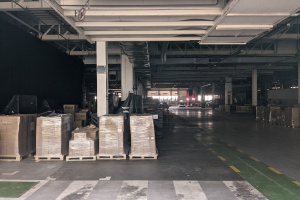 Großes Lager mit zahlreichen Kartons auf Paletten, gestützt von Säulen, unter Deckenbeleuchtung und einem Schild, das ein Flottenmanagementsystem anzeigt.