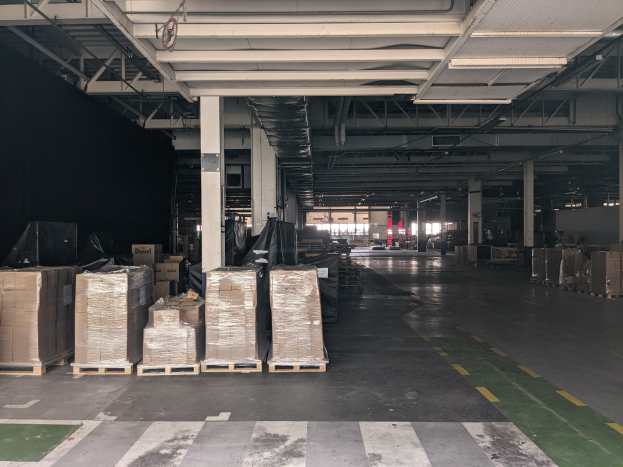 Großes Lager mit zahlreichen Kartons auf Paletten, gestützt von Säulen, unter Deckenbeleuchtung und einem Schild, das ein Flottenmanagementsystem anzeigt.