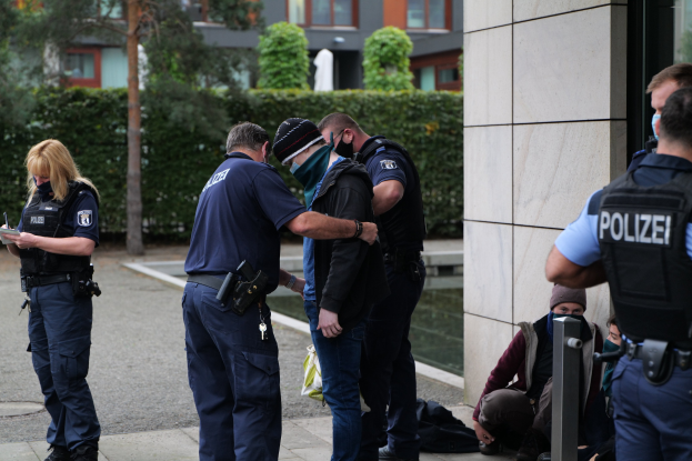 Polizisten vor einem Gebäude mit einer Frau, die ein Buch und einen Stift hält, während andere in der Nähe sitzen und stehen, mit Bäumen und Gebäuden im Hintergrund.