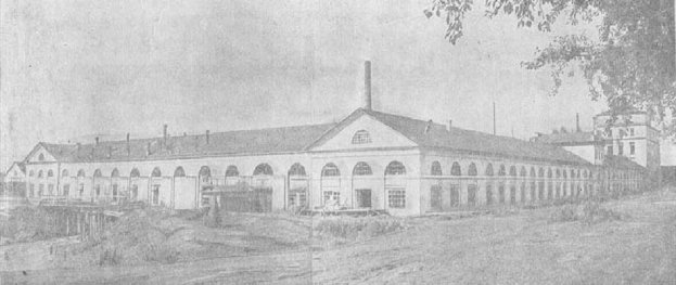 Schwarz-weiß-Foto eines großen, mehrfenstrigen Gebäudes, das von Grün umgeben ist und unter einem klaren Himmel steht, mit Text unten, der lautet: "1892 – das erste Fabrikgebäude in der Stadt".
