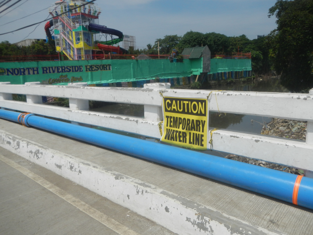 Eine Brücke mit einem "Vorsicht temporäre Wasserleitung"-Schild, Geländern und einem Hintergrund mit Bäumen, Gebäuden, einer Wasserrutsche und einem bewölkten Himmel.