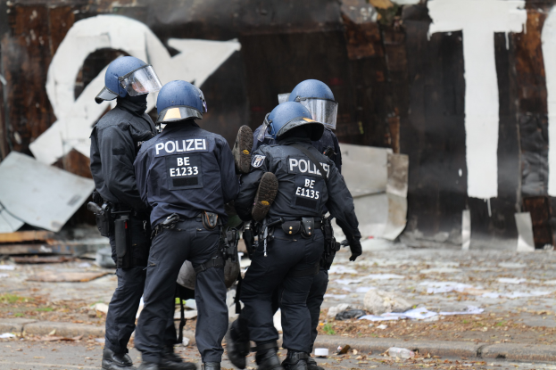 Polizeibeamte in schwarzen Uniformen und blauen Helmen stehen vor einem Gebäude mit Graffiti, umgeben von verstreuten Papieren und Gegenständen.