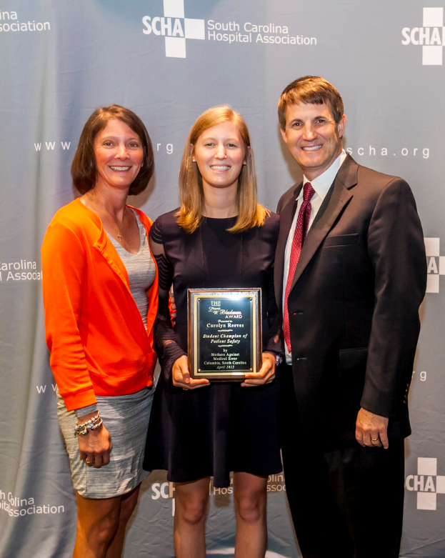 Drei Menschen, zwei Frauen und ein Mann, die zusammenstehen, wobei eine Frau einen Preis hält, vor einem grauen Hintergrund.